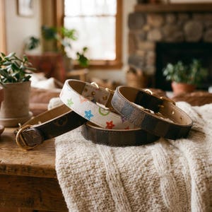 May include: Three dog collars with gold-colored hardware rest on a wooden table covered with a cream-colored blanket. One collar is white with colorful star shapes, another is brown with a repeating logo pattern, and the third is a darker brown with a checkered pattern.