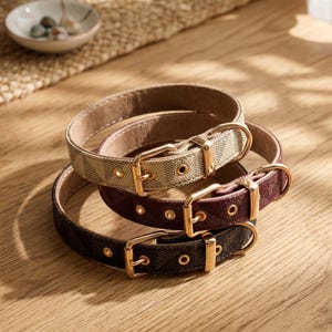 May include: Three pet collars stacked together, featuring a repeating geometric pattern in beige, burgundy, and dark brown. Each collar has a gold-toned metal buckle, a matching metal loop, and reinforced eyelets for adjustment, with a light-colored lining.