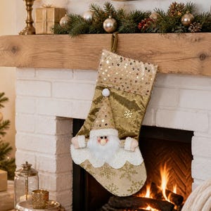 May include: A gold and cream Christmas stocking featuring a plush Santa Claus figure with a white beard and a sequined hat. The stocking has a cuff covered in gold sequins, a metallic gold body with snowflake patterns, and a small gold snowflake.