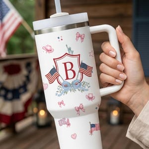 May include: A white insulated tumbler with a handle and straw featuring a patriotic pattern of American flags, pink bows, cowboy hats, hearts, and a central red shield emblem with the letter B. The design includes the text USA in pink letters.