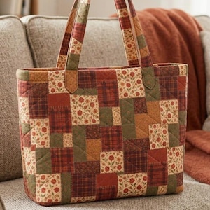 May include: A quilted tote bag featuring a patchwork design of rectangular fabric pieces in shades of rust orange, olive green, and brown. The pattern includes floral prints and plaid textures with visible stitching throughout the exterior.