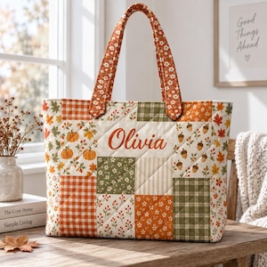 May include: A quilted tote bag featuring a patchwork design of autumn-themed patterns including pumpkins, acorns, leaves, and gingham in orange, green, and white. The name Olivia is embroidered in orange script on the center panel.