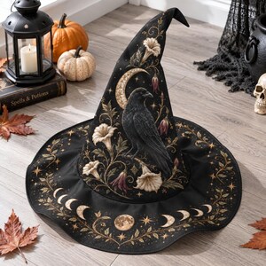 May include: A black pointed hat featuring a printed design of a raven perched on a branch, a crescent moon, and floral motifs. The brim is decorated with a sequence of moon phases and stars in gold and cream tones against the dark fabric.