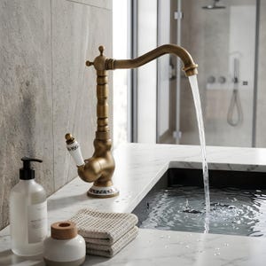 May include: An antique-style brass faucet with a curved spout, pouring water into a dark sink. The faucet has a decorative handle and base with floral patterns. A bottle of soap, a small wooden container, and folded towels are on the marble countertop.