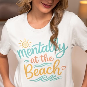 Puede incluir: Camiseta blanca con la frase "mentally at the beach" en turquesa, amarillo y naranja. El diseño incluye un sol, olas y un corazón. Prenda de vestir informal con temática playera.