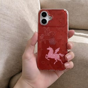 May include: A red phone case with a clear border, featuring a pink Pegasus design. The case has a glossy finish and is decorated with small star and heart shapes. The phone case is held in a hand.