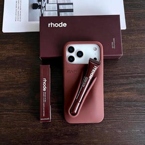 May include: A dark maroon smartphone case with a built-in holder for a tube of lip tint, accompanied by a matching rectangular box and a slim tube of lip tint, all featuring the white rhode logo and text printed on the surfaces.