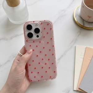 Könnte beinhalten: Eine transparente Handyhülle mit rosa Hintergrund und roten Punkten. Die Hülle wird in einer Hand mit einem Perlenring gehalten. Die Handyhülle hat einen Kameraausschnitt und Tastenabdeckungen.