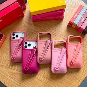 May include: A collection of silicone phone cases in various shades of pink, each featuring a moulded pocket designed to hold a small tube of rhode lip treatment. The cases are branded with the word rhode and are displayed alongside matching boxes.