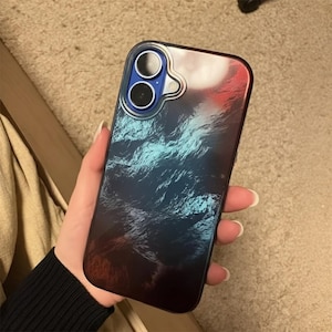 Op de afbeelding: Een smartphonehoesje met een gradiëntontwerp. De hoes gaat over van rood naar blauw, met een gestructureerd, golfachtig patroon. De cameralenzen zijn zichtbaar door een heldere, beschermende ring.