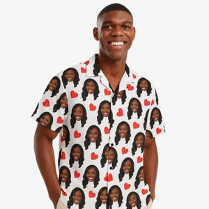 May include: A short-sleeved button-down shirt with a white base colour featuring a repeating pattern of red hearts and a smiling person's face with long dark hair. The shirt has a classic collar and a front button closure with dark buttons.