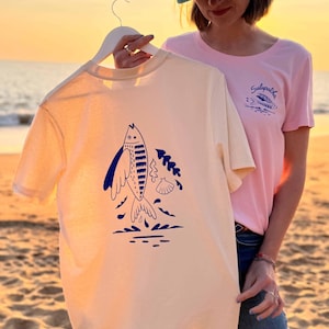 Peut inclure: Un t-shirt beige clair avec une illustration bleue d'un poisson, d'algues et d'un coquillage. Le t-shirt est tenu par un cintre blanc. Un t-shirt rose avec le mot "Galapatba" est visible en arrière-plan.