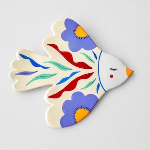 Könnte beinhalten: Eine dekorative Keramikschale in Vogelform mit cremefarbenem Grund. Der Vogel hat blaue, rote und grüne stilisierte Blumen- und Blattmotive. Der Kopf des Vogels ist weiß mit geschlossenem Auge, rotem Punkt und orangem Schnabel.