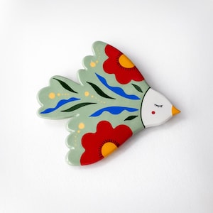 May include: A decorative bird figurine with a light green body and red floral accents. The bird has a white head with a closed eye, a red cheek, and a yellow beak. Blue and black details complete the design.