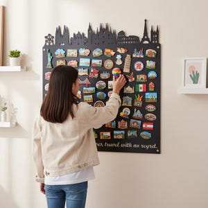 Può includere: Una lavagna magnetica nera con una silhouette di skyline in alto, piena di colorati magneti souvenir a tema viaggio. Una persona sta posizionando un magnete a forma di mongolfiera sulla lavagna. Il testo in basso recita: "...travel with no regret."
