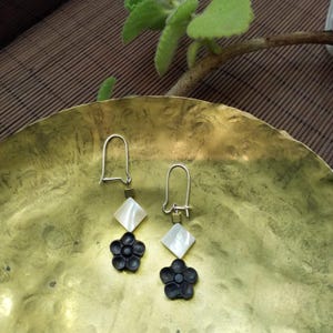 May include: A pair of silver earrings with a distinctive design. Each earring has a white, diamond-shaped element, a small square accent, and a black flower-shaped charm. The earrings are presented on a gold-coloured, textured surface.