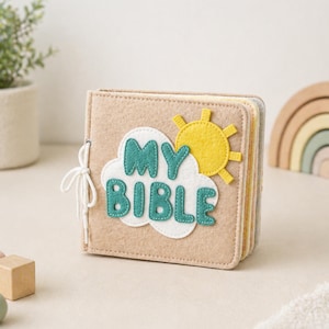 May include: A beige felt book with the words "MY BIBLE" in teal letters on a white cloud, and a yellow sun. The book is tied with a white string. The pages are visible and have different colors.