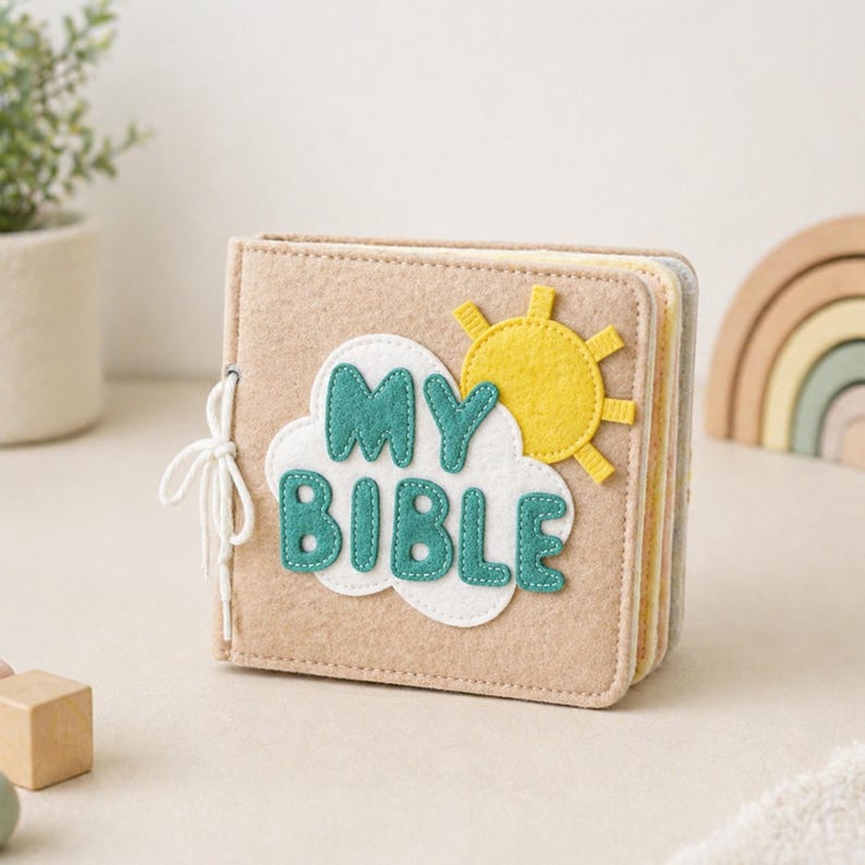 May include: A beige felt book with the words "MY BIBLE" in teal letters on a white cloud, and a yellow sun. The book is tied with a white string. The pages are visible and have different colors.