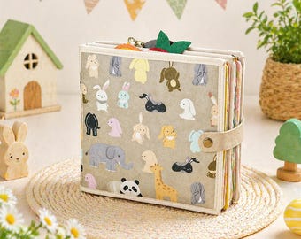 May include: A beige journal with a variety of animal illustrations, including rabbits, elephants, and giraffes. The journal has a beige suede strap closure and a green felt strawberry charm. The journal is sitting on a woven mat.