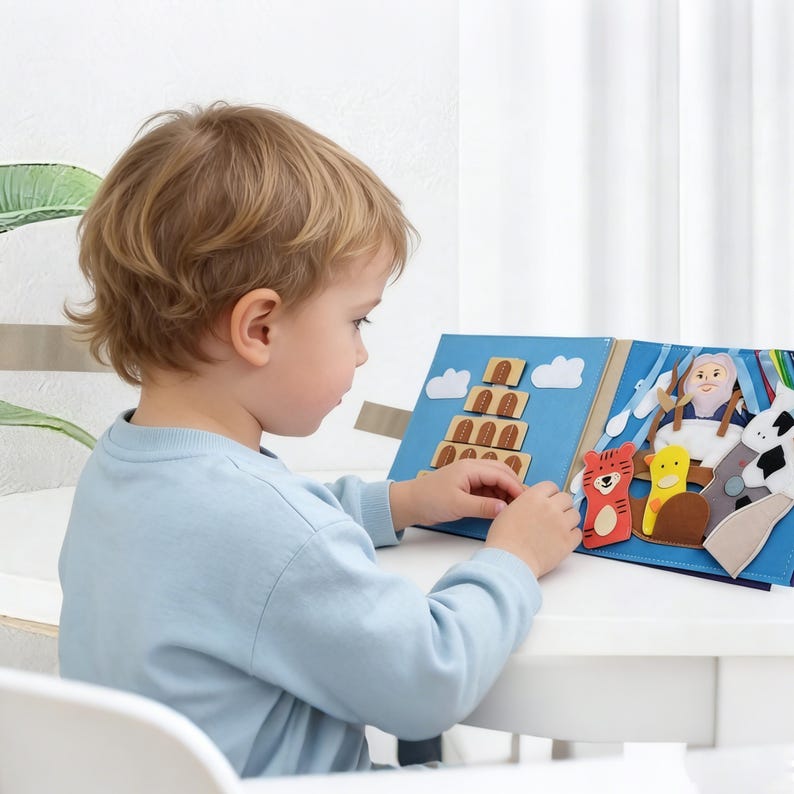 May include: A young child is looking at a blue felt book with various felt cutouts. The book features a tower, clouds, and animal figures. The child is wearing a light blue long-sleeve shirt and is sitting at a white table.