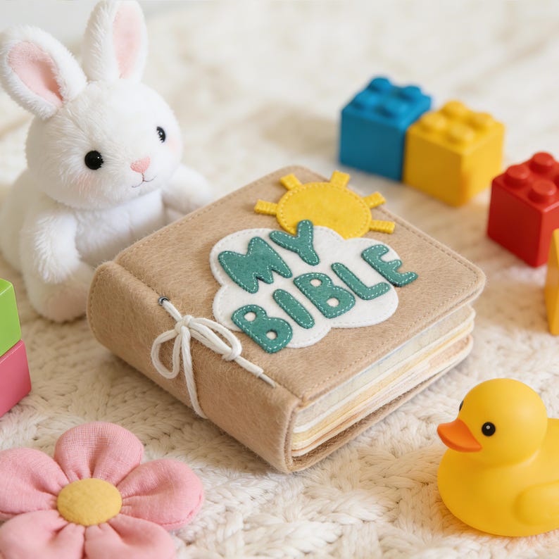 May include: A tan felt book with the words "MY BIBLE" in teal letters and a white cloud design. A yellow sun is above the cloud. A white stuffed bunny, colorful blocks, a pink flower, and a yellow rubber duck are nearby.