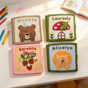 Può includere: Quattro libri in feltro colorati con disegni ricamati. Un libro presenta un orso con il nome "Olivitte", un altro un fungo con "Lauraoly", un terzo con fragole e "Sarahlis", e l'ultimo con un leone e "Alicelyo".