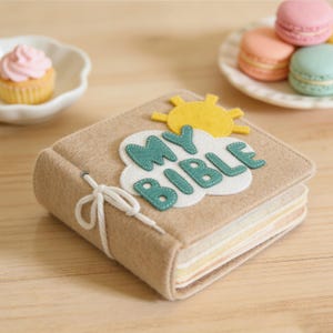 May include: A tan felt book with the words "MY BIBLE" appliqued on the cover, along with a yellow sun and white cloud. The book is tied with a white string. A cupcake and macarons are in the background.