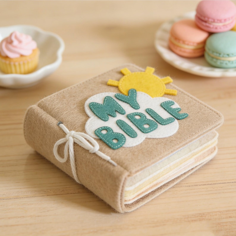 May include: A tan felt book with the words "MY BIBLE" appliqued on the cover, along with a yellow sun and white cloud. The book is tied with a white string. A cupcake and macarons are in the background.