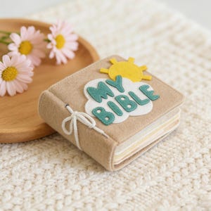 May include: A beige felt book with the words "MY BIBLE" appliqued on the cover. The letters are teal with white stitching, set against a white cloud. A yellow sun is above the cloud. The book is tied with a white string.