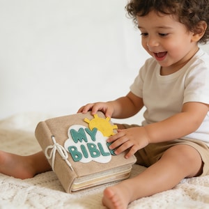 May include: A beige felt book with the words "MY BIBLE" on the cover, featuring a cloud and sun design. The book is tied with a white string. The book is being held by a child.