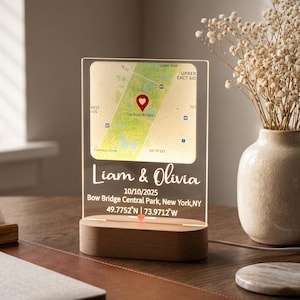 Puede incluir: Una lámpara de mapa acrílica personalizada con base de madera. El mapa resalta una ubicación con un marcador de corazón, con los nombres "Liam & Olivia" y la fecha 10/10/2025. Un jarrón con flores secas está cerca.