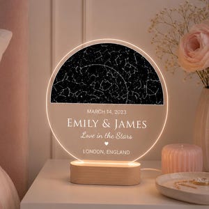 以下が含まれることがあります： 星図デザインのパーソナライズされたナイトライト。ライトには「EMILY & JAMES Love in the Stars」と「LONDON, ENGLAND」の文字が。木製のベースに置かれ、白いベッドサイドテーブルの上にあります。