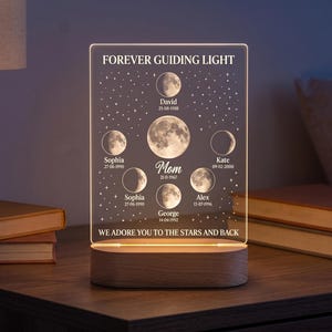 May include: A personalized LED night light with the text "FOREVER GUIDING LIGHT" and the phrase "WE ADORE YOU TO THE STARS AND BACK". The clear acrylic panel features moon phases and names with dates, set on a wooden base. The light is on a wooden table.