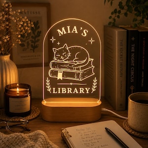 Może przedstawiać: Łukowata lampka nocna LED z akrylu z drewnianą podstawą, z grawerowaną ilustracją kota śpiącego na stosie książek. Napis głosi MIA'S LIBRARY, z małymi akcentami w kształcie gwiazdek i liści. Lampka emituje ciepłe, złociste światło.