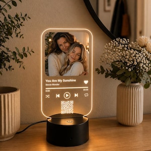 Può includere: Una luce notturna personalizzata in acrilico con una foto e i dettagli della canzone. La luce presenta una foto di due persone, il titolo della canzone "You Are My Sunshine" di Stevie Wonder e un codice QR. La base è nera e la luce ha un bagliore caldo.