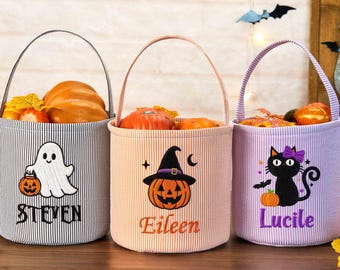 May include: Three cylindrical fabric buckets with handles, each featuring a Halloween-themed embroidered design and a name. The left bucket is grey-striped with a ghost and the name Steven. The center is peach with a pumpkin and Eileen. The right is purple with a black cat and Lucile.