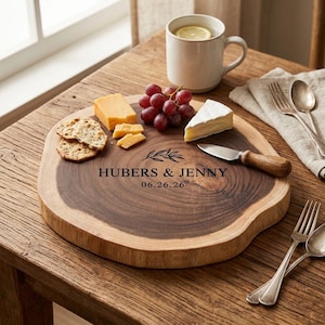 Puede incluir: Una tabla de servir redonda de madera con bordes de corteza natural, grabada con el texto HUBERS & JENNY y 06.26.26, junto a una ilustración de hoja. La tabla contiene galletas, queso, uvas y un cuchillo con mango de madera.