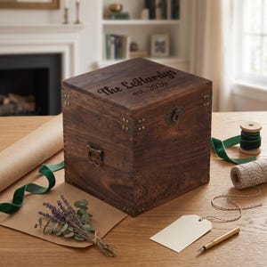 May include: A dark brown wooden box with a hinged lid, featuring metal corner reinforcements and a front latch. The top is engraved with the text "The LeHardys est. 2026". The box has side handles and a visible wood grain texture throughout the surface.