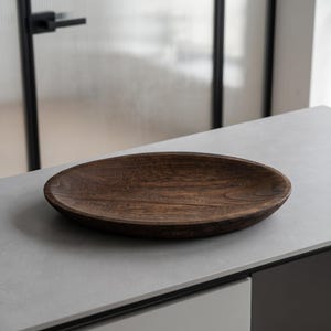 Pode incluir: Uma tigela oval de madeira com acabamento castanho escuro e um padrão visível de grão de madeira natural. A tigela apresenta um interior liso e raso com uma borda curva, destacando a textura orgânica e os tons profundos do material de madeira.
