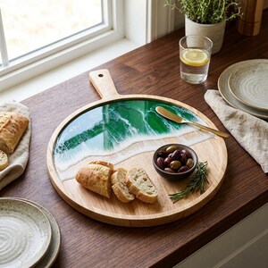 Könnte beinhalten: Ein rundes Servierbrett aus Holz mit Griff, das eine dekorative Harzeinlage aufweist, die eine grüne Meereswelle mit weißem Schaum nachahmt. Das Brett enthält geschnittenes Brot, eine kleine Schale mit dunklen Oliven und ein goldenes Streichmesser.