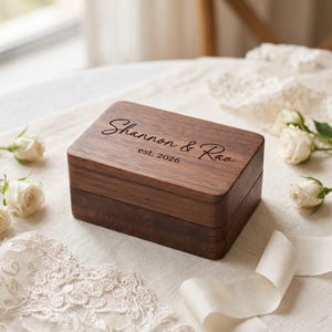 May include: A rectangular dark brown wooden keepsake box with a smooth finish. The lid is engraved with the text "Shannon & Rao" in a cursive script font, followed by the text "est. 2026" in a simple, clean, sans-serif typeface.