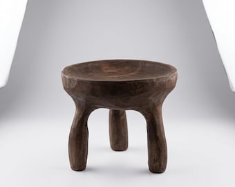 May include: A dark brown wooden stool featuring a circular, slightly concave seat and three thick, hand-carved legs. The surface of the wood displays a textured, uneven finish with visible chisel marks throughout the entire structure of the stool.