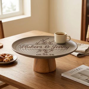 Puede incluir: Un soporte para pasteles redondo de cerámica gris con base de madera. La superficie presenta un diseño botánico grabado con láser, el texto "Hubers & Jenny" y la fecha "06.26.26". Una pequeña taza de cerámica a juego descansa sobre el plato.