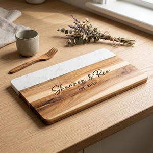 May include: A rectangular serving board featuring a combination of natural wood and white marble. The wood section is engraved with the text Shannon & Rao and est. 2026. A small ceramic cup, a wooden fork, and a dried lavender sprig are nearby.