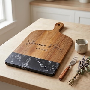 May include: A rectangular wooden serving board with a handle and a black marble accent at the bottom. The wood is engraved with the text Shannon & Rao and est. 2026 in a script and sans-serif font. The board has a natural grain finish.