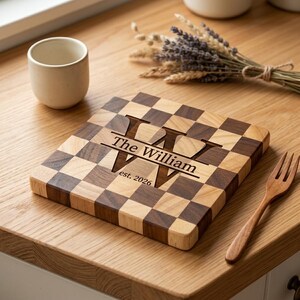 May include: A square wooden cutting board featuring a checkerboard pattern of light and dark wood squares. The center is engraved with a large letter W, the text The William, and the date est. 2026. The board has a smooth, flat surface.