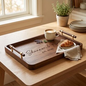 May include: A rectangular dark wood serving tray with two horizontal wooden handles featuring gold-tone metal accents. The tray is engraved with the text "Shannon & Ray" and "est. 2026". It holds a ceramic mug, a small plate with a croissant, and a linen napkin.