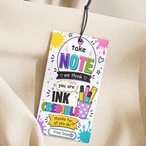 Puede incluir: Una colorida etiqueta de regalo con las palabras "take NOTE" en una burbuja. La etiqueta también dice "INK CREDIBLE" y "thanks for all you do!" con una taza de lunares morados que contiene bolígrafos y una regla. La etiqueta está sujeta a una cuerda negra.