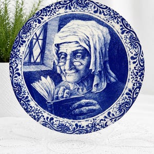 May include: A blue and white decorative plate with a detailed illustration of an elderly person reading a book. The plate has a floral border and is displayed on a clear stand. The image is reminiscent of Delftware.