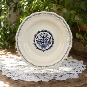 Peut inclure: Une assiette en céramique blanche avec un bord festonné et un motif floral bleu au centre. L'assiette a une bordure bleue délicate et est présentée sur un napperon en dentelle blanche, idéale pour servir des aliments.